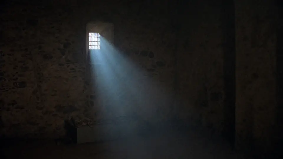 Celda antigua de piedra con un rayo de luz que entra por la ventana y un rollo sobre la banca